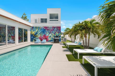 Your modern tropical oasis awaits with a vibrant mural, heated pool, gas grill, pool loungers and seating area framed by swaying palms—perfect for sunny relaxation in style!