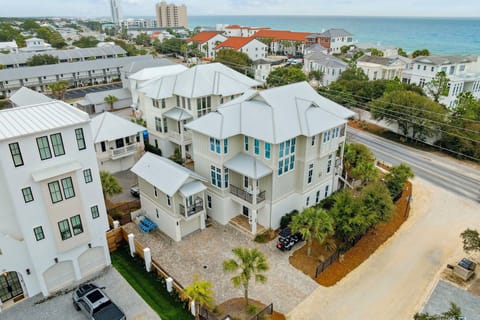 Aerial view of a stunning beachside property featuring modern coastal architecture with white metal roofing, surrounded by lush landscaping in a vibrant neighborhood near the ocean.