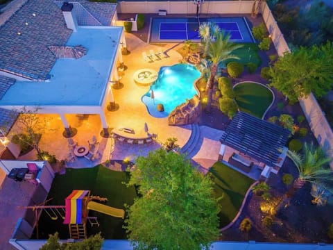 Aerial view of a resort-style backyard featuring a freeform pool with rock waterfall, tennis court, outdoor dining patio, playground, and lush tropical landscaping illuminated at twilight.