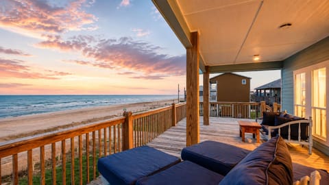 Oceanfront deck lounge at sunset with cushioned seating, railing, covered balcony, and sweeping beach views.