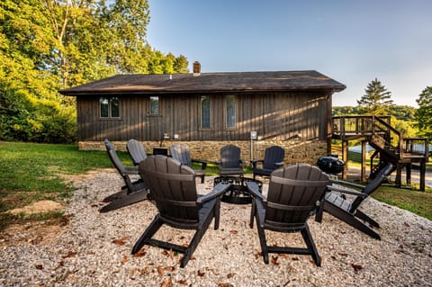 Eight black Adirondack chairs encircle a metal fire pit on white gravel beside a rustic wood-sided house.