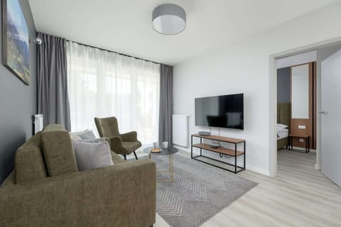 A modern living room with a soft green armchair and a cozy gray sofa facing a TV. Large windows fill the space with natural light.