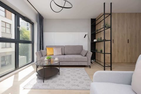 A cozy living room space with a comfortable gray sofa and access to a sunny balcony. Natural light fills the room, enhancing the light wood and neutral decor.