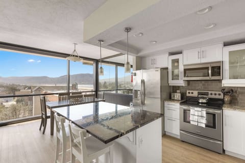 Your open-concept culinary and dining haven with a view! This bright space features a modern kitchen island, a dining table, and expansive windows showcasing stunning coastal and town scenery.