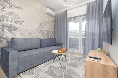 A cozy living room with a grey sofa, patterned rug, and coffee table, perfect for relaxing. Large windows with curtains allow plenty of natural light.