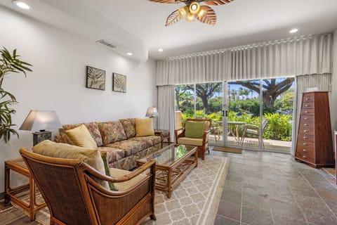 Relax in your bright tropical living room with plush seating, unique leaf-motif ceiling fan, and sliding doors opening to lush garden views perfect for unwinding.