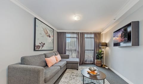 A bright, contemporary living space featuring an L-shaped grey sectional sofa, a round coffee table on an area rug, and a wall-mounted TV.