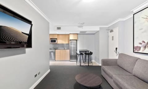 A wide shot from the living area showing a grey sofa, a round coffee table, a wall-mounted TV, and the kitchen in the background.