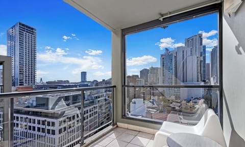 A bright, glass-enclosed balcony corner with white tiling, offering a wide look out onto the city high-rises under a clear blue sky.