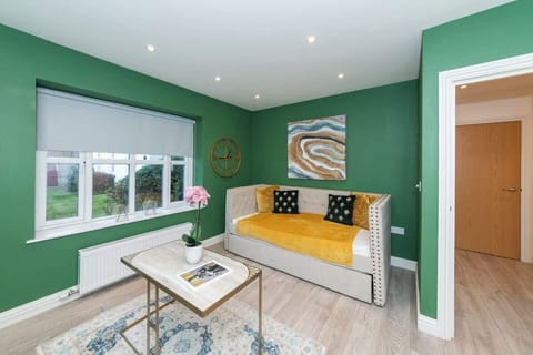 A modern living room with green walls, a cream sofa with yellow and black cushions, a large abstract painting, a window with a white blind, a coffee table with a plant, and light wood flooring.
