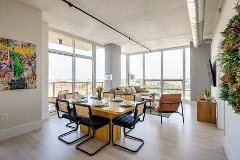 Open-concept dining and living area with large windows. The dining space features a wooden table with six chairs, and the living room has a sectional sofa, chairs, and a 65` TV. Decor includes a colorful wall art piece and a vertical plant display.