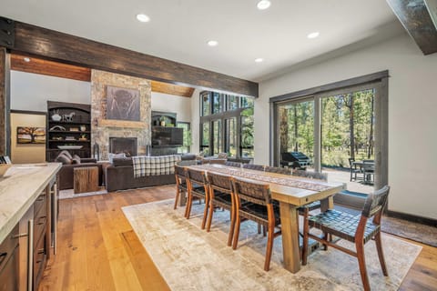 Gather around the spacious dining table where natural light pours through expansive windows, framing serene forest views. High ceilings with rustic wood beams create an airy, welcoming space perfect for memorable meals together.