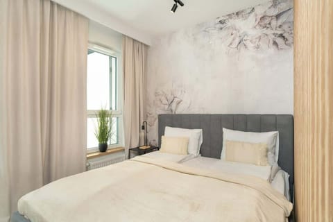 A cozy corner with a double bed, beige and gray bedding, decorative wall art, and a large window letting in natural light.