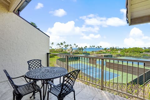 A peaceful private balcony with comfortable seating offering scenic views of nearby tennis courts, tropical palms, and the distant ocean under blue skies.