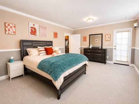 Spacious and stylish bedroom with plush carpeting, a cozy cream bed, vibrant artwork, and a soft teal throw blanket. Warm neutral tones, modern furnishings, and thoughtful décor create a relaxing retreat perfect for unwinding after a day of exploring