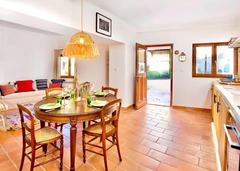 The open-plan kitchen and dining area features a charming rustic table set for four. Terracotta tile floors and exposed beams create a warm, inviting atmosphere, while French doors open to the serene outdoor terrace. A unique woven pendant light adds an a