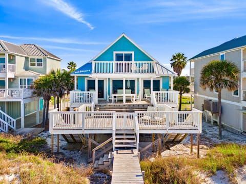 Charming turquoise beach house on stilts with multiple decks and boardwalk, surrounded by coastal palms in a sunny waterfront community.