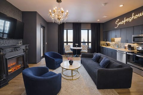 Enjoy a modern, open-plan living space with vibrant blue furniture, a dark blue sofa, and a stylish kitchen with stainless steel appliances and pendant lighting. A chandelier adds elegance to the cohesive design.
