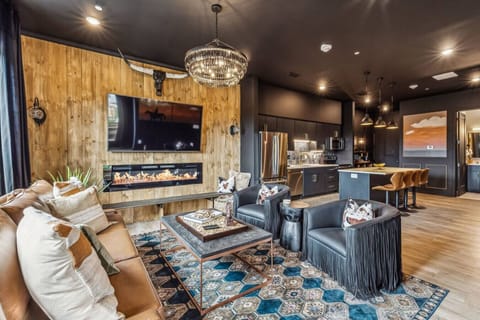 Western-themed living area with a rustic, wood-paneled accent wall featuring a mounted TV and an electric fireplace beneath it. The room is furnished with a leather sofa, black leather chairs with fringe detailing.