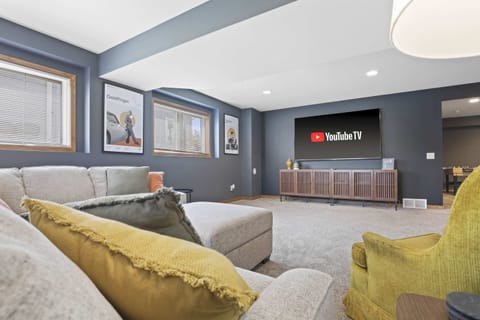 Lower level game room with large sectional sofa, 80" smart TV, arcade game, foosball, and kids play room.