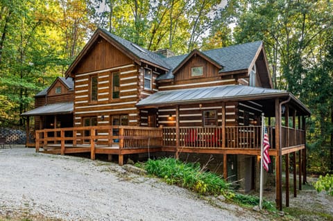 Log cabin exterior with wraparound porch, red porch swing, and wood railing surrounded by tall trees.