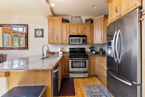 A modern kitchen with wooden cabinets, stainless steel appliances, and a marble countertop. A clock is on the wall above a window with a view.