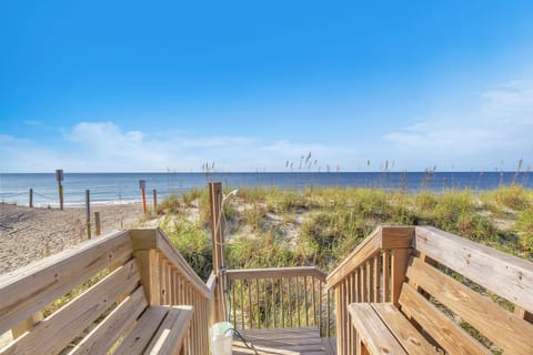 Oceanfront deck with built-in benches, wood railings, outdoor shower, dune grass, beach access path, wide ocean views, sandy shore, and blue sky.
