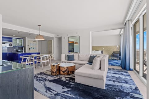 Open‑concept luxury condo with L‑shaped sofa, full kitchen, dining area and floor‑to‑ceiling windows opening to a huge private balcony with oversized turf and relaxing ocean view.