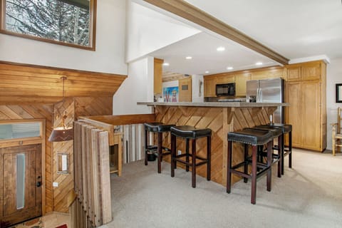 An open kitchen with a wooden bar counter and three bar stools, adjacent to a staircase with wooden railings, and a wooden front door partially visible in the foreground.