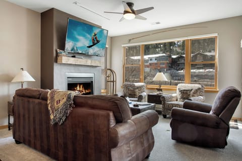 A living room with a fireplace, wall-mounted TV, and a large window. There are two brown sofas and two patterned armchairs arranged around a coffee table. A ceiling fan is on the white ceiling.