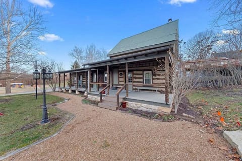 The larger historic log cabin welcomes guests with a long covered porch, landscaped gravel paths, and mature trees.