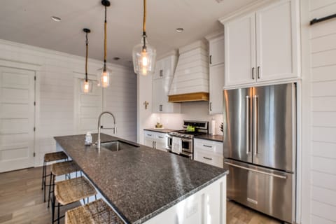 Kitchen island with pendant lighting, gas range, and stainless s - Designer Kitchen – Gas Range, Pendant Lighting & Storage Galore – A closer look at the heart of this St. Simons vacation rental kitchen, featuring a deep island with seating, dramatic lighting, a Keurig + drip coffee setup, and sleek stainless steel finishes.
