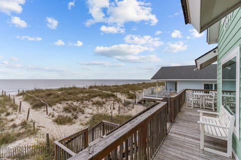 Oak Island, NC deck with white bench, dining table, wood railings, dune walkway, sea oats, beach views, neighboring homes, and wide Atlantic Ocean scenery.
