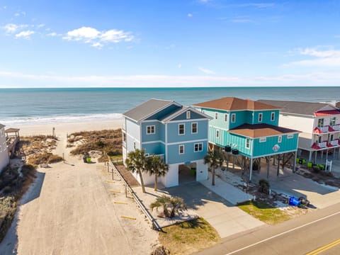 Bright blue raised home with palm trees, white gravel landscaping, concrete driveway, neighboring teal beach house, and ocean views just steps from the sandy shoreline.