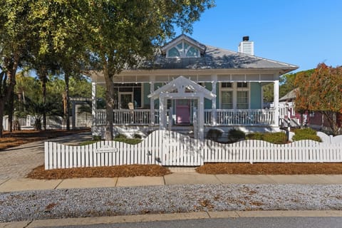 Charming coastal cottage with wraparound porch and picket fence, nestled among mature trees in a peaceful neighborhood setting.
