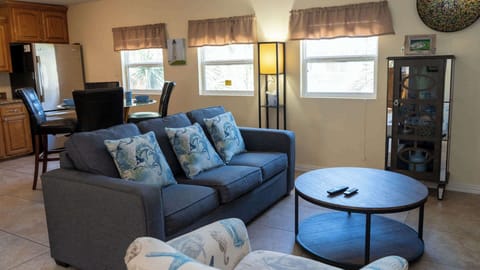 Cozy Nautical Lounge & Kitchenette with Tropical Vibes
Relax on deep gray sofa with blue crab pillows, facing glass-top coffee table. Sunny windows frame palms; adjacent kitchen boasts wood cabinets, fridge, dinette. Beige walls, lamp glow, wall art.