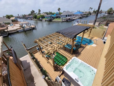 Waterfront deck features a wooden pergola with shaded lounge chairs, a mini putting green, and bubbling hot tub.