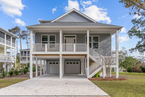 Gray beach rental with covered porch, white railings, double garage, wide driveway, side stairs, green lawn, and blue sky.