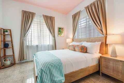 A bedroom with a queen-size bed dressed in white linens and a light blue throw. Two windows with beige curtains allow natural light. A wooden nightstand and soft lighting frame the bed.