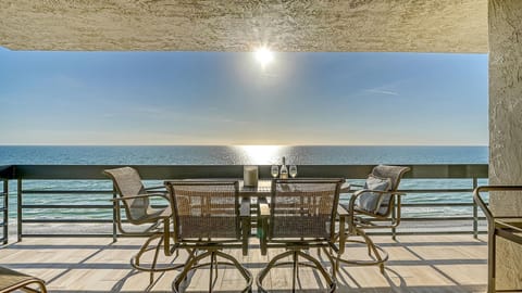 Sunlight sparkles on the ocean beyond the balcony’s metal railing and four mesh swivel chairs around a glass-top table.
