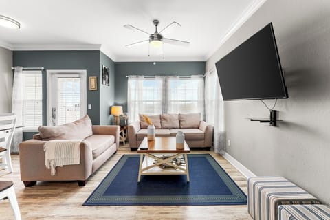This cozy living room welcomes you with warm natural light streaming through the large windows, highlighting the inviting ambiance of this space. The plush sofas, paired with a stylish coffee table, provide a perfect spot for relaxation. A sleek wall-mounted TV ensures entertainment is just a click away, while the modern ceiling fan adds both comfort and style. The navy blue ar...