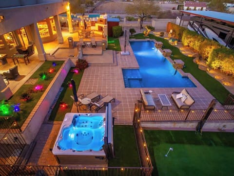 An aerial view of the beautifully lit backyard reveals a modern pool, hot tub, and spacious patio with colorful garden lighting perfect for evening entertaining and relaxation.