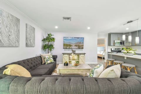 Elegant Largo pool home near Indian rocks beach living room showcasing plush tufted sectional, coastal artwork, and modern farmhouse décor. Open-concept design with kitchen access and premium entertainment setup. 