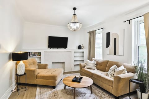 The living room features a beige sofa and chaise, round wood coffee table, TV, decorative fireplace, built-in shelving, windows, chandelier, and area rug.