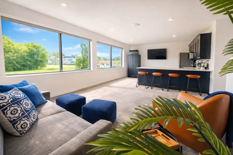A spacious lounge area bathed in natural light from wall-to-wall windows, featuring a cozy sectional, stylish poufs, and modern seating. Perfect for relaxing or entertaining with scenic views and seamless access to the kitchen and bar.