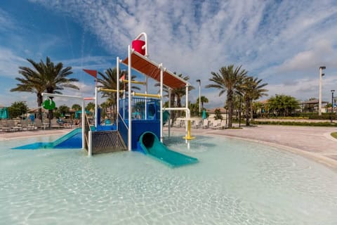 A vibrant, zero-entry splash park area featuring a colorful playground structure with slides in a clear, shallow pool, surrounded by palm trees.