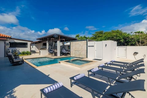 An outdoor space with a pool, hot tub, lounge chairs with striped towels, and a shaded pergola with swing chairs and a grill. White walls and greenery surround the area under a clear blue sky.