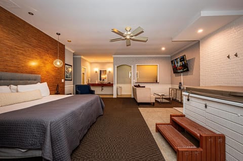 Suite features a king bed with gray quilt, a white brick hot tub with wooden steps, and a wall-mounted TV.