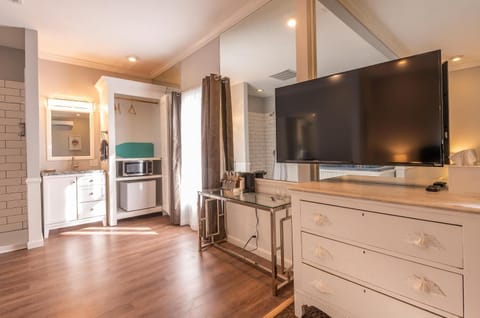 Flat-screen TV mounted above a white dresser with shell drawer pulls beside a glass-top desk and open closet.