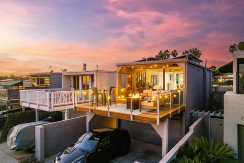 The house features a spacious outdoor deck lit by cozy string lights, providing a warm ambiance. The open sliding doors reveal a well-lit and inviting interior, seamlessly blending indoor and outdoor living spaces.